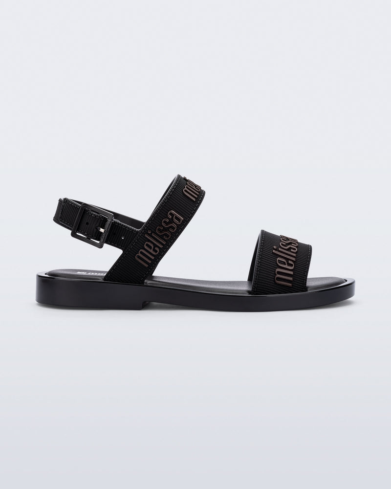 Side view of the Melissa Mar Sandal with Melissa logo embossed into the straps. There are two embossed straps that go across the foot; 1 across the toes and the other across the main part of the foot. The main foot strap has an ankle strap attached with a buckle closure. The straps ,buckle and sole of the shoe is black. The Melissa logo on the straps has a black metallic finish. . The buckle placement is on the outside of the ankle strap. 
