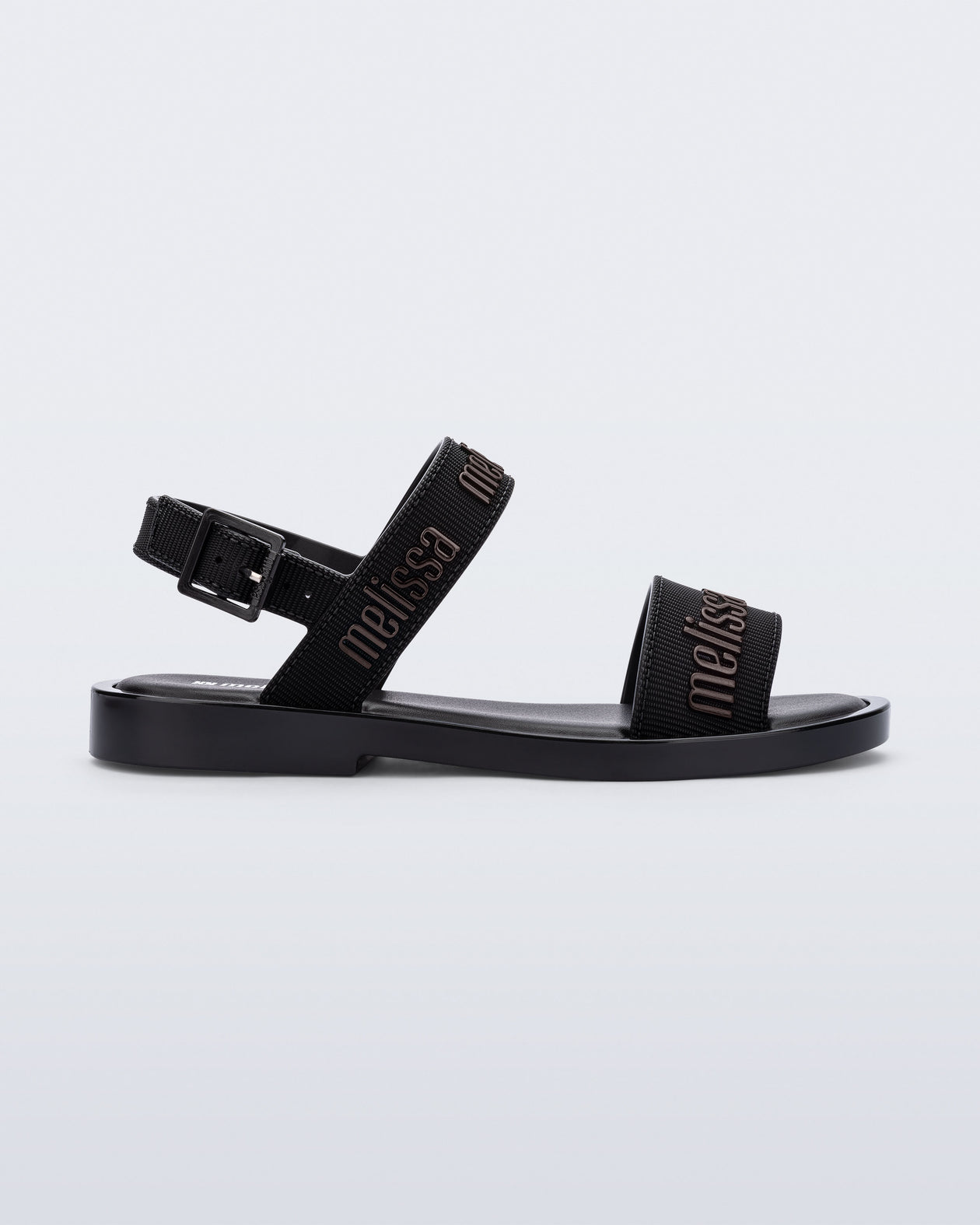 Side view of the Melissa Mar Sandal with Melissa logo embossed into the straps. There are two embossed straps that go across the foot; 1 across the toes and the other across the main part of the foot. The main foot strap has an ankle strap attached with a buckle closure. The straps ,buckle and sole of the shoe is black. The Melissa logo on the straps has a black metallic finish. . The buckle placement is on the outside of the ankle strap. 