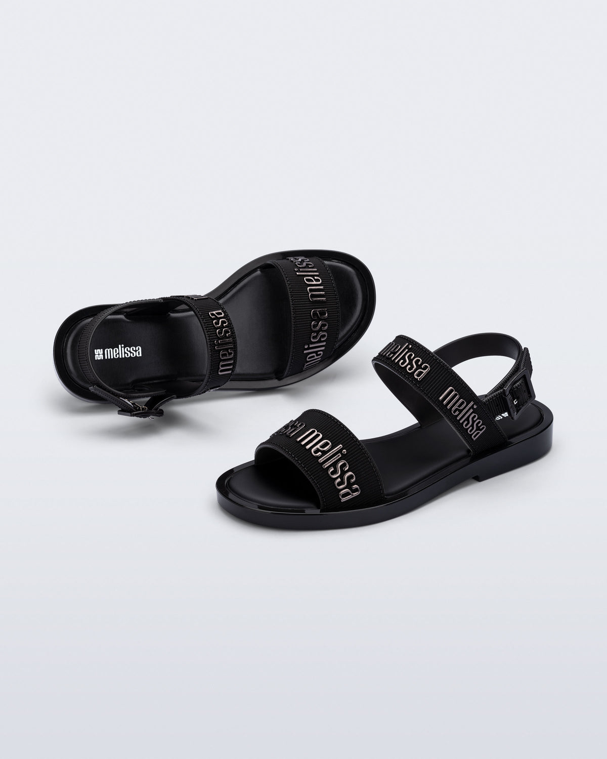 Angled view of a pair of Melissa Mar Sandals with Melissa logo embossed into the straps. There are two embossed straps that go across the foot; 1 across the toes and the other across the main part of the foot. The main foot strap has an ankle strap attached with a buckle closure. The straps ,buckle and sole of the shoe is black. The Melissa logo on the straps has a black metallic finish. The buckle placement is on the outside of the ankle strap. 