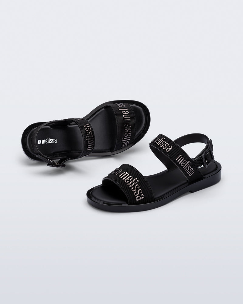 Angled view of a pair of Melissa Mar Sandals with Melissa logo embossed into the straps. There are two embossed straps that go across the foot; 1 across the toes and the other across the main part of the foot. The main foot strap has an ankle strap attached with a buckle closure. The straps ,buckle and sole of the shoe is black. The Melissa logo on the straps has a black metallic finish. The buckle placement is on the outside of the ankle strap. 