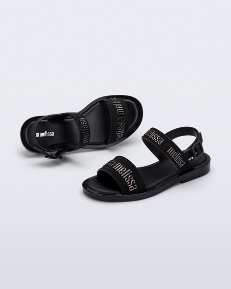 Angled view of a pair of Melissa Mar Sandals with Melissa logo embossed into the straps. There are two embossed straps that go across the foot; 1 across the toes and the other across the main part of the foot. The main foot strap has an ankle strap attached with a buckle closure. The straps ,buckle and sole of the shoe is black. The Melissa logo on the straps has a black metallic finish. The buckle placement is on the outside of the ankle strap. 