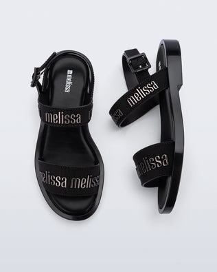 Top and side view of a pair of Melissa Mar Sandals with Melissa logo embossed into the straps. There are two embossed straps that go across the foot; 1 across the toes and the other across the main part of the foot. The main foot strap has an ankle strap attached with a buckle closure. The straps ,buckle and sole of the shoe is black. The Melissa logo on the straps has a black metallic finish. The buckle placement is on the outside of the ankle strap. 