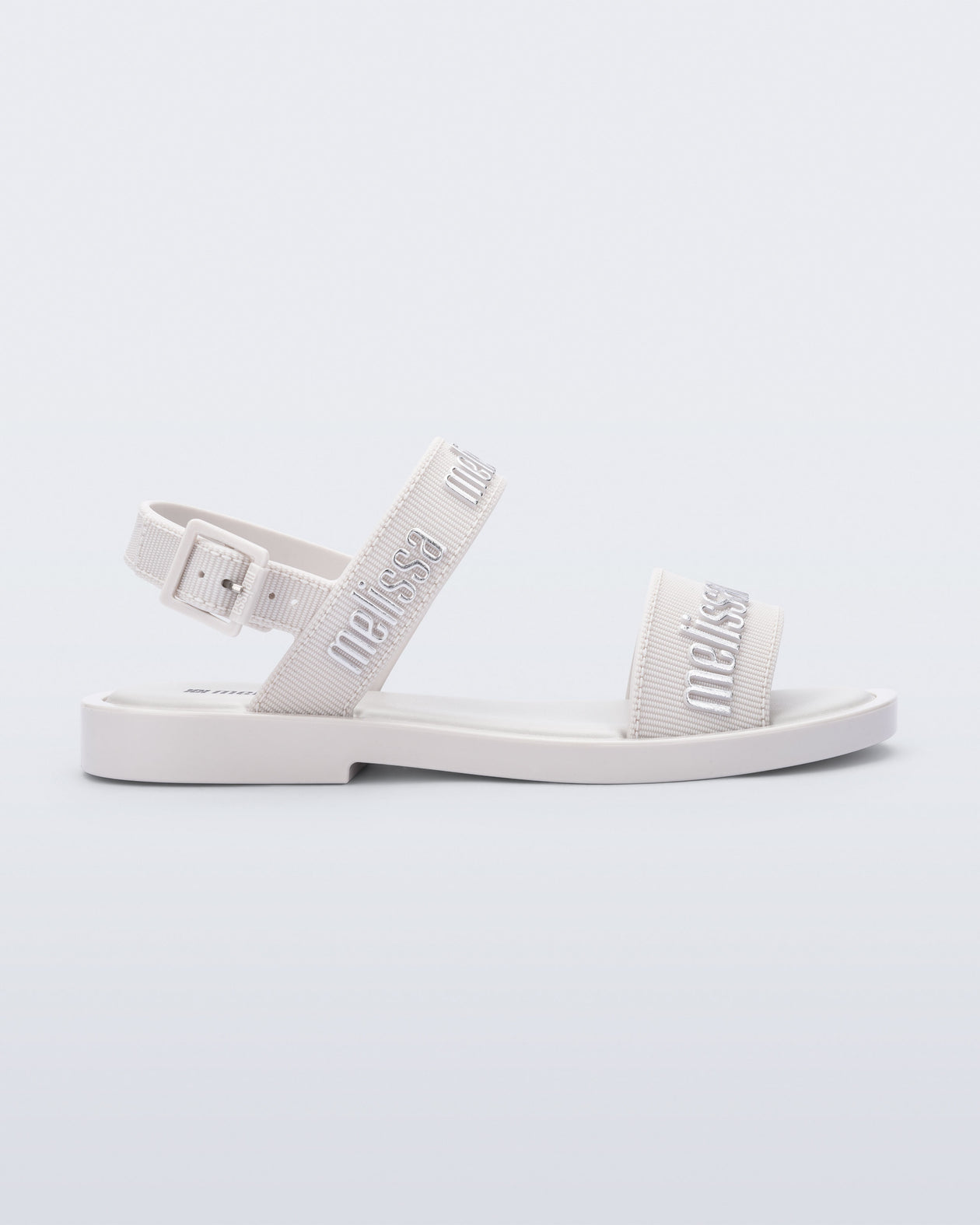 Side view of the Melissa Mar Sandal with Melissa logo embossed into the straps. There are two embossed straps that go across the foot; 1 across the toes and the other across the main part of the foot. The main foot strap has an ankle strap attached with a buckle closure. The straps ,buckle and sole of the shoe is white. The Melissa logo on the straps has a silver metallic finish. . The buckle placement is on the outside of the ankle strap. 
