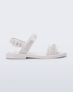 Side view of the Melissa Mar Sandal with Melissa logo embossed into the straps. There are two embossed straps that go across the foot; 1 across the toes and the other across the main part of the foot. The main foot strap has an ankle strap attached with a buckle closure. The straps ,buckle and sole of the shoe is white. The Melissa logo on the straps has a silver metallic finish. . The buckle placement is on the outside of the ankle strap. 