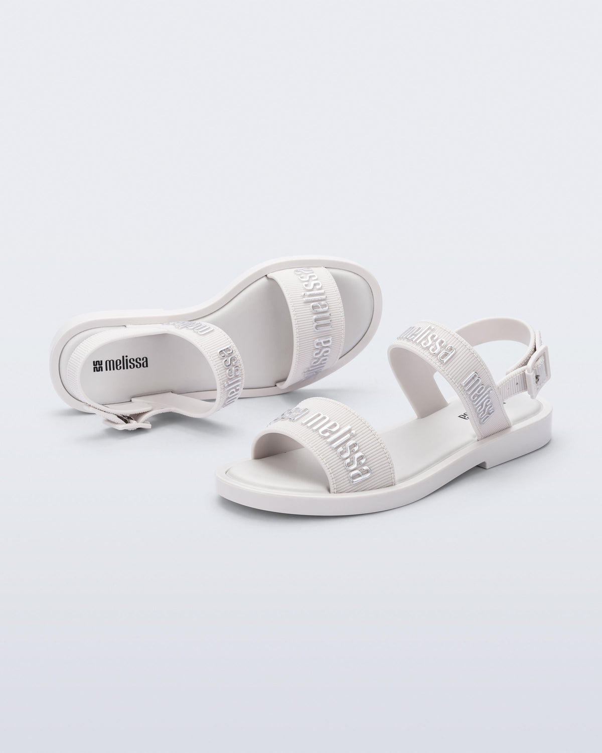 Angled view of a pair of Melissa Mar Sandals with Melissa logo embossed into the straps. There are two embossed straps that go across the foot; 1 across the toes and the other across the main part of the foot. The main foot strap has an ankle strap attached with a buckle closure. The straps ,buckle and sole of the shoe is white. The Melissa logo on the straps has a silver metallic finish. The buckle placement is on the outside of the ankle strap. 