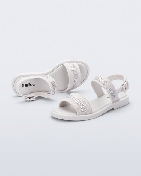 Angled view of a pair of Melissa Mar Sandals with Melissa logo embossed into the straps. There are two embossed straps that go across the foot; 1 across the toes and the other across the main part of the foot. The main foot strap has an ankle strap attached with a buckle closure. The straps ,buckle and sole of the shoe is white. The Melissa logo on the straps has a silver metallic finish. The buckle placement is on the outside of the ankle strap. 