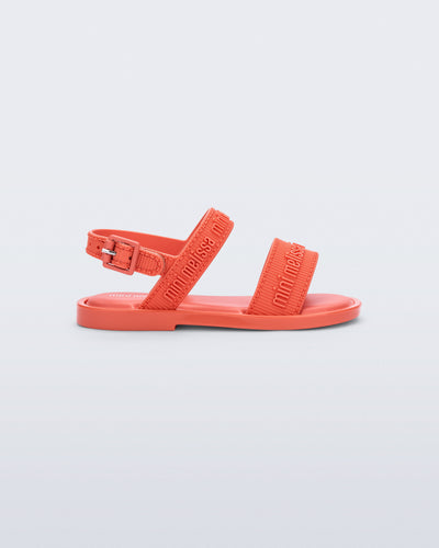 Side view of the Mini Melissa Mar Sandal with Mini Melissa logo embossed into the straps. There are two embossed straps that go across the foot; 1 across the toes and the other across the main part of the foot. The main foot strap has an ankle strap attached with a buckle closure. The buckle placement is on the outside of the ankle strap. The color of the shoe is orange.