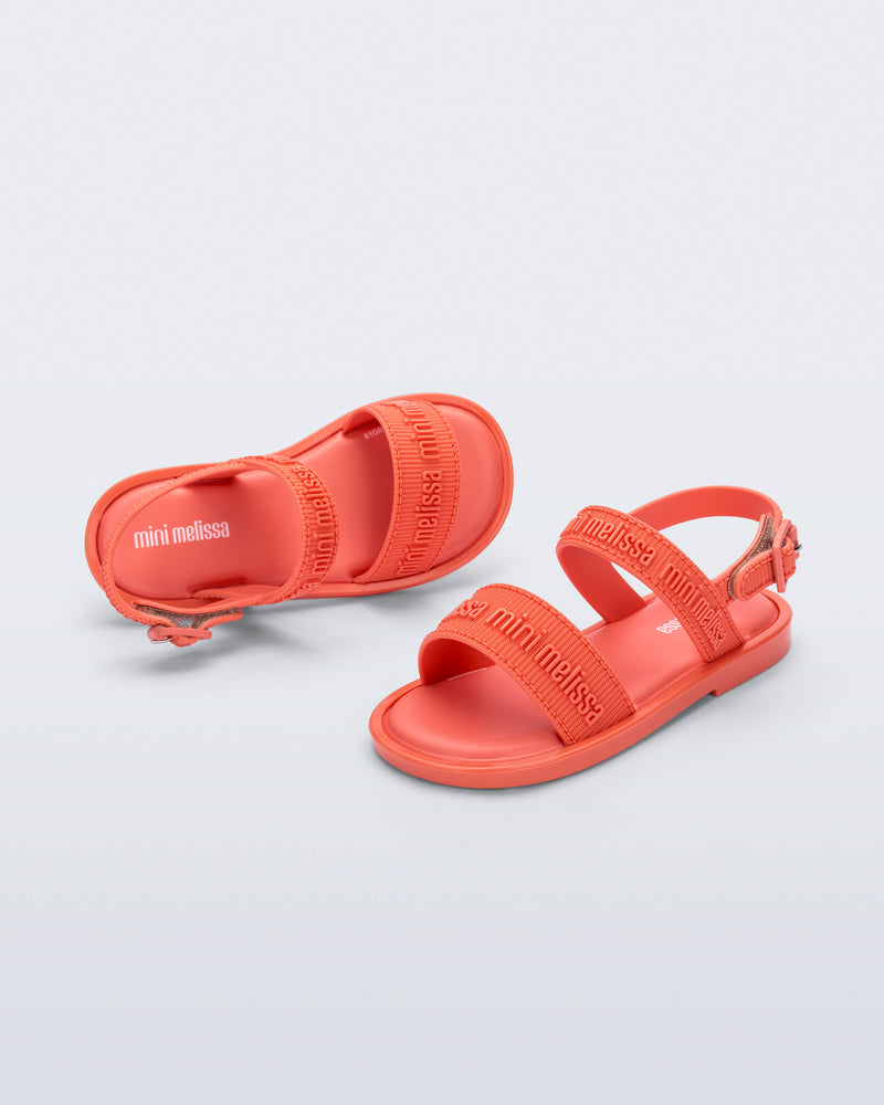 Angled view of a pair of Mini Melissa Mar Sandals with Mini Melissa logo embossed into the straps. There are two embossed straps that go across the foot; 1 across the toes and the other across the main part of the foot. The main foot strap has an ankle strap attached with a buckle closure. The buckle placement is on the outside of the ankle strap. The color of the shoe is orange.