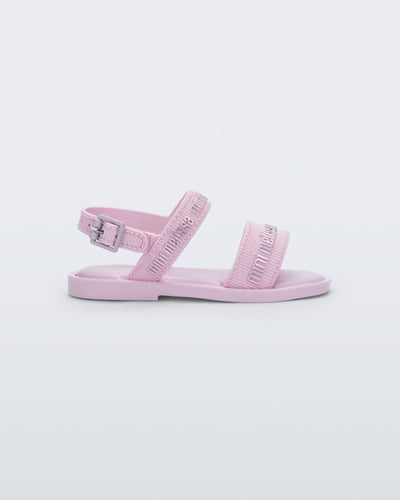 Side view of the Mini Melissa Mar Sandal with Mini Melissa logo embossed into the straps. There are two embossed straps that go across the foot; 1 across the toes and the other across the main part of the foot. The main foot strap has an ankle strap attached with a buckle closure. The straps ,buckle and sole of the shoe is pink. The Melissa logo on the straps has a silver metallic finish. . The buckle placement is on the outside of the ankle strap. 