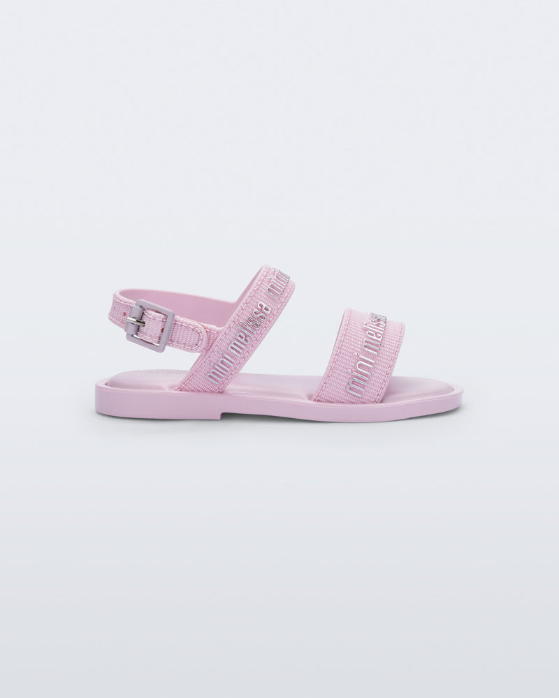 Side view of the Mini Melissa Mar Sandal with Mini Melissa logo embossed into the straps. There are two embossed straps that go across the foot; 1 across the toes and the other across the main part of the foot. The main foot strap has an ankle strap attached with a buckle closure. The straps ,buckle and sole of the shoe is pink. The Melissa logo on the straps has a silver metallic finish. . The buckle placement is on the outside of the ankle strap. 