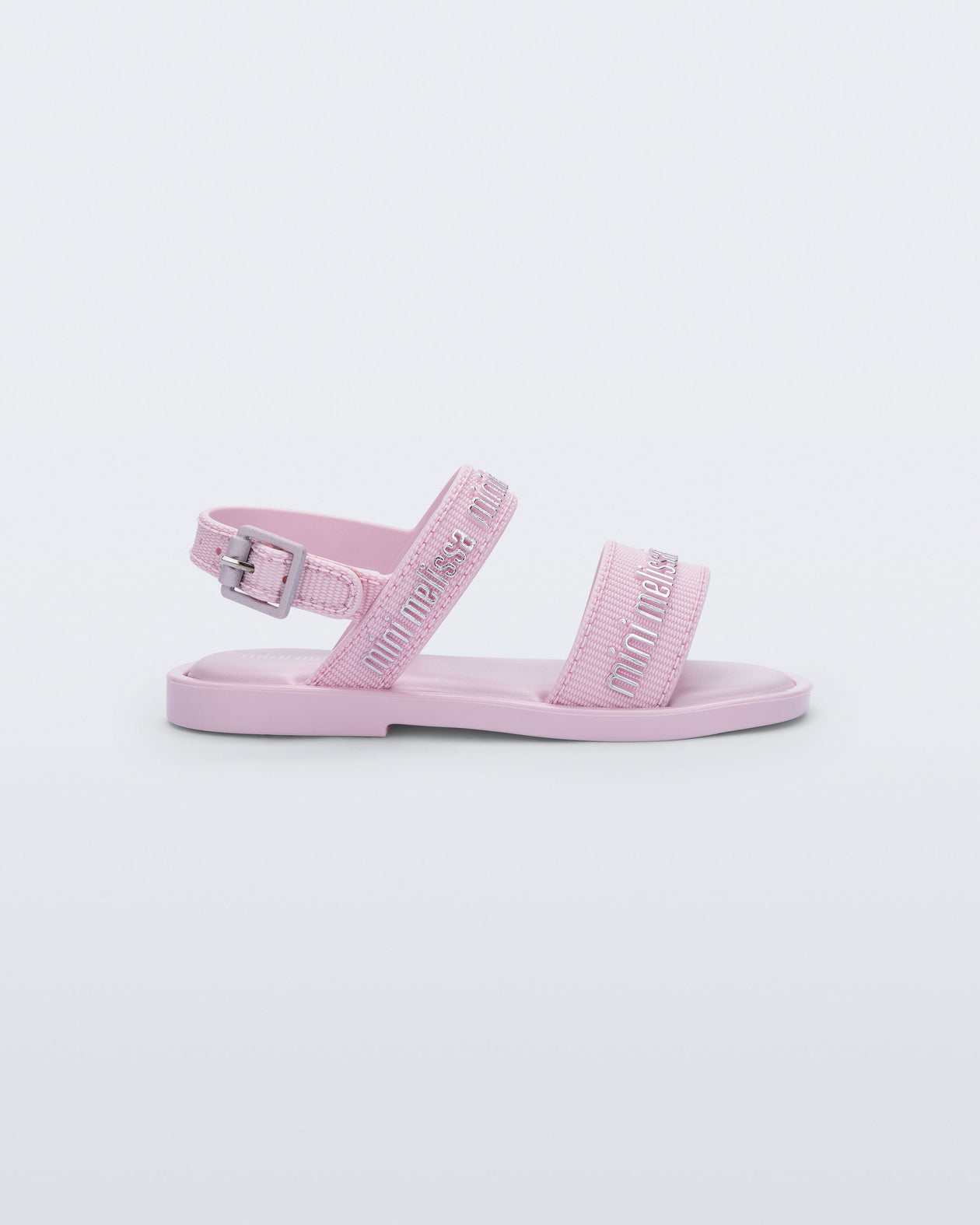 Side view of the Mini Melissa Mar Sandal with Mini Melissa logo embossed into the straps. There are two embossed straps that go across the foot; 1 across the toes and the other across the main part of the foot. The main foot strap has an ankle strap attached with a buckle closure. The straps ,buckle and sole of the shoe is pink. The Melissa logo on the straps has a silver metallic finish. . The buckle placement is on the outside of the ankle strap. 
