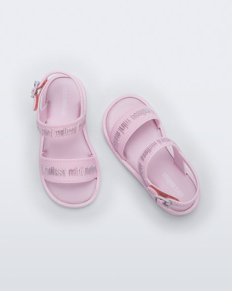 Top view of a pair of Mini Melissa Mar Sandals with Mini Melissa logo embossed into the straps. There are two embossed straps that go across the foot; 1 across the toes and the other across the main part of the foot. The main foot strap has an ankle strap attached with a buckle closure. The straps ,buckle and sole of the shoe is pink. The Melissa logo on the straps has a silver metallic finish. The buckle placement is on the outside of the ankle strap. 