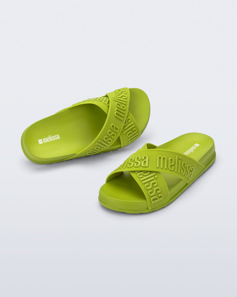 Angled view of a pair of Melissa Cozy slide sandals. The slide has two cross straps with Melissa logo embossed onto the straps. The color of the shoe is a lime green. 