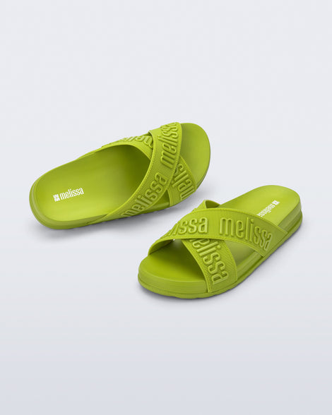 Angled view of a pair of Melissa Cozy slide sandals. The slide has two cross straps with Melissa logo embossed onto the straps. The color of the shoe is a lime green. 