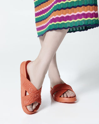 Close up view of a model's legs posing in a pair of Melissa Cozy slide sandals. The slide has two cross straps with Melissa logo embossed onto the straps. The color of the shoe is orange