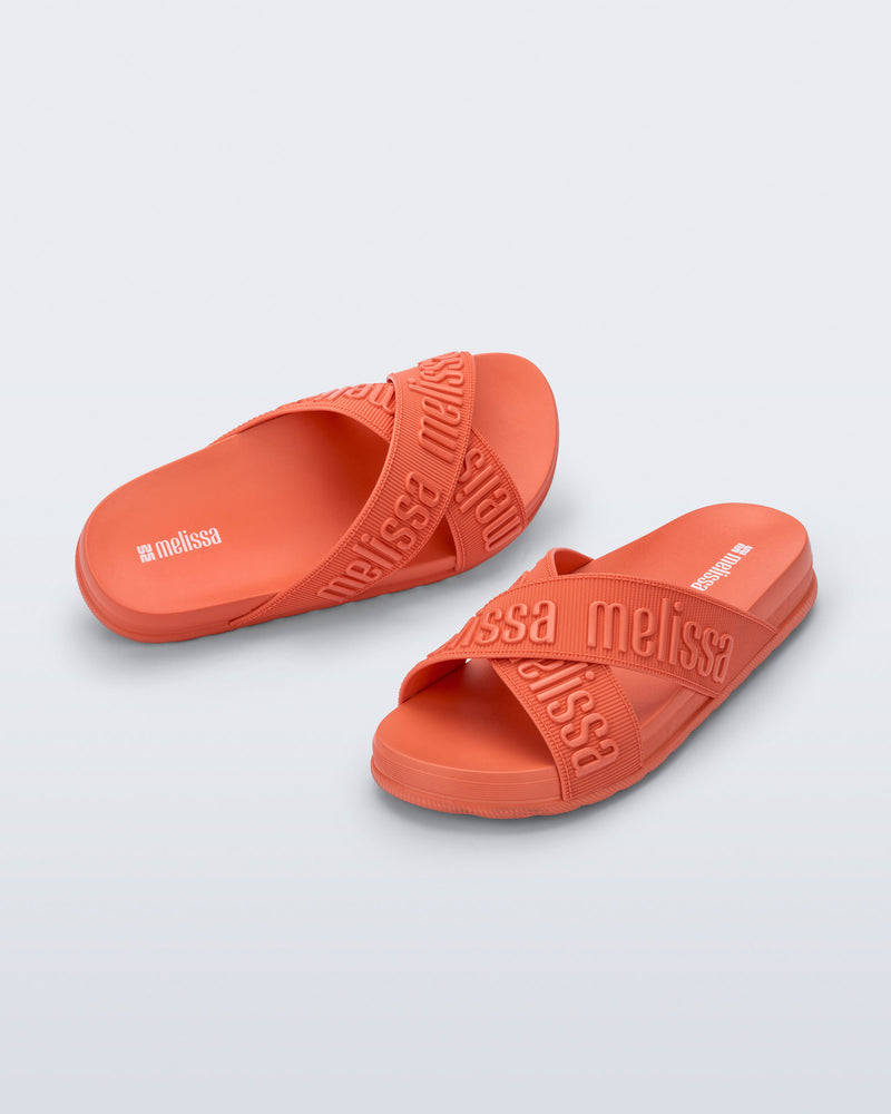 Angled view of a pair of Melissa Cozy slide sandals. The slide has two cross straps with Melissa logo embossed onto the straps. The color of the shoe is orange.