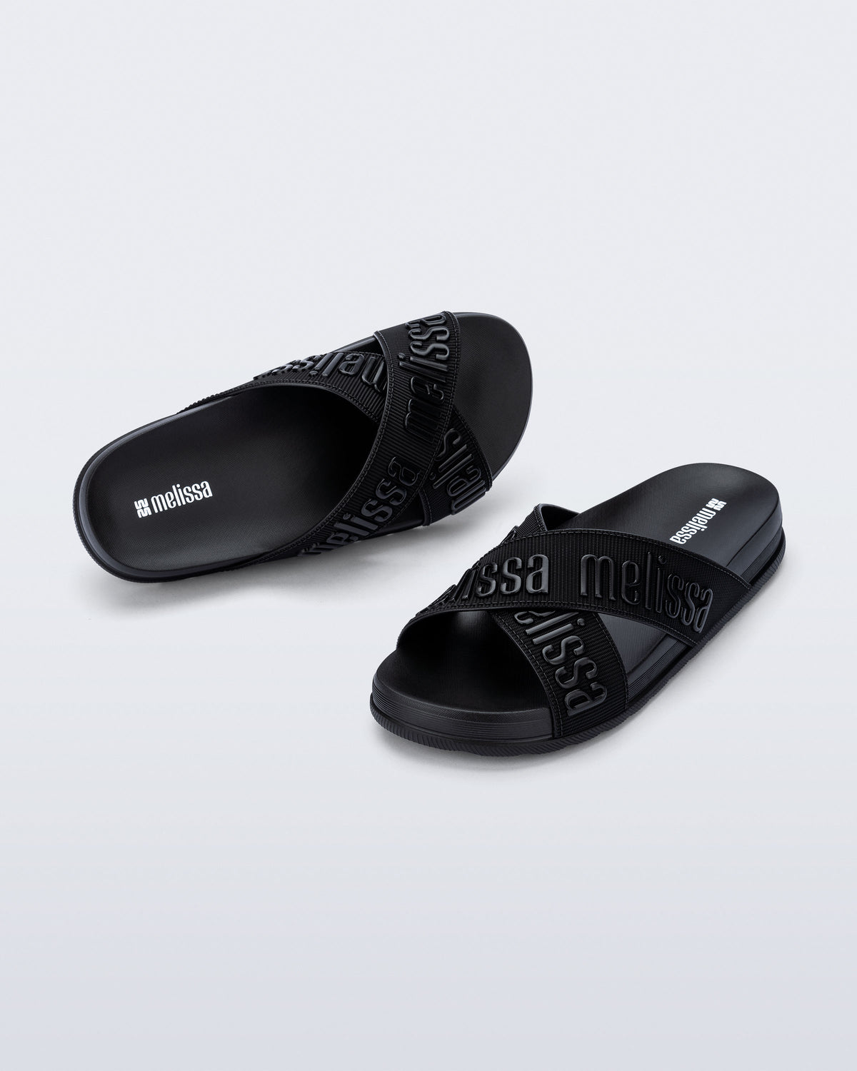 Angled view of a pair of Melissa Cozy slide sandals. The slide has two cross straps with Melissa logo embossed onto the straps. The color of the shoe is black.
