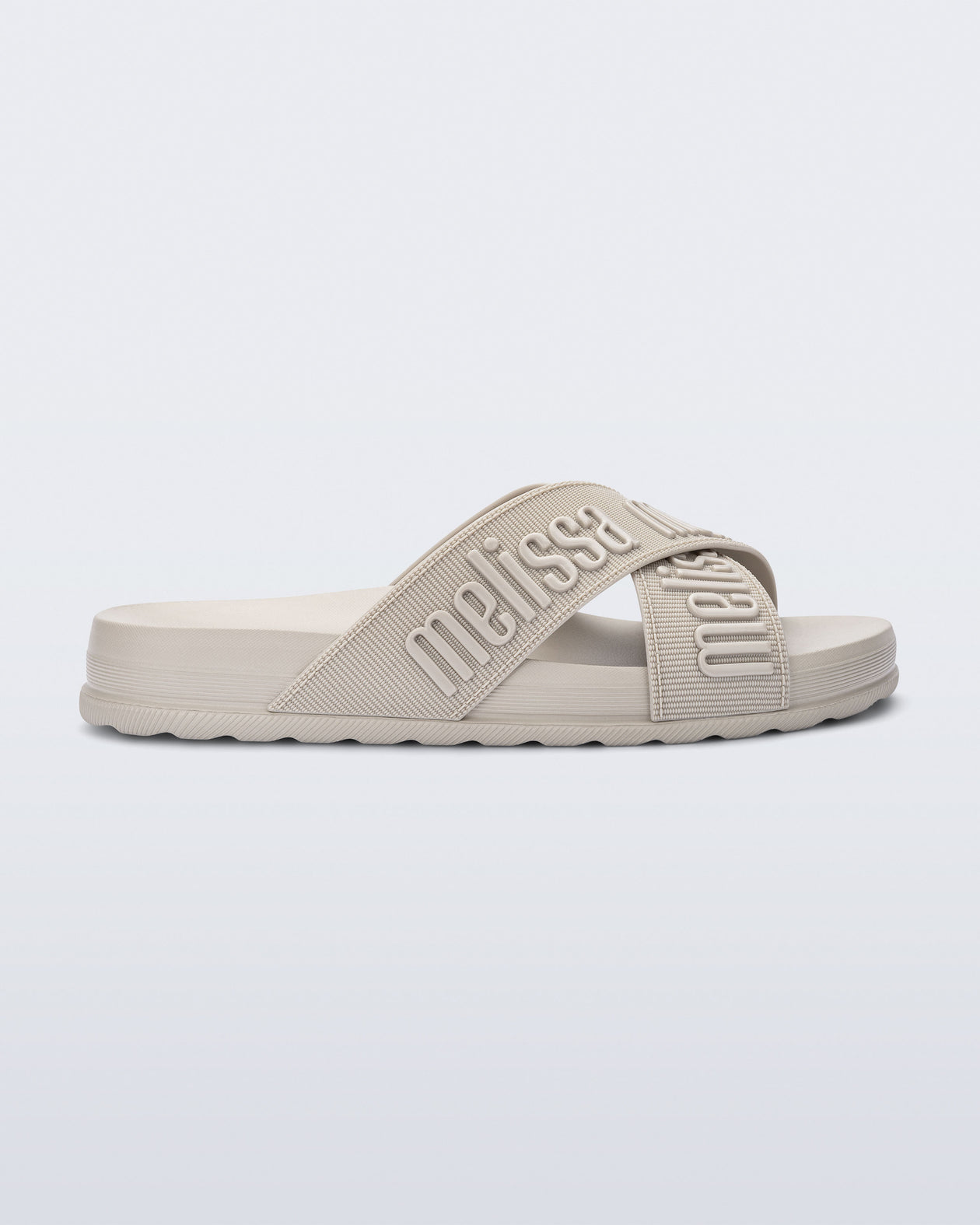 Side view of the Melissa Cozy slide sandals. The slide has two cross straps with Melissa logo embossed onto the straps. The color of the shoe is beige. 