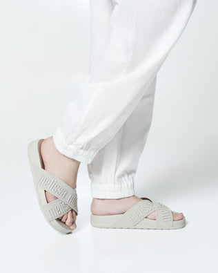 Close up view of a model's legs posing in a pair of Melissa Cozy slide sandals. The slide has two cross straps with Melissa logo embossed onto the straps. The color of the shoe is beige