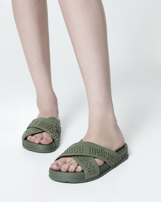 Close up view of a model's legs posing in a pair of Melissa Cozy slide sandals. The slide has two cross straps with Melissa logo embossed onto the straps. The color of the shoe is a dark green. 