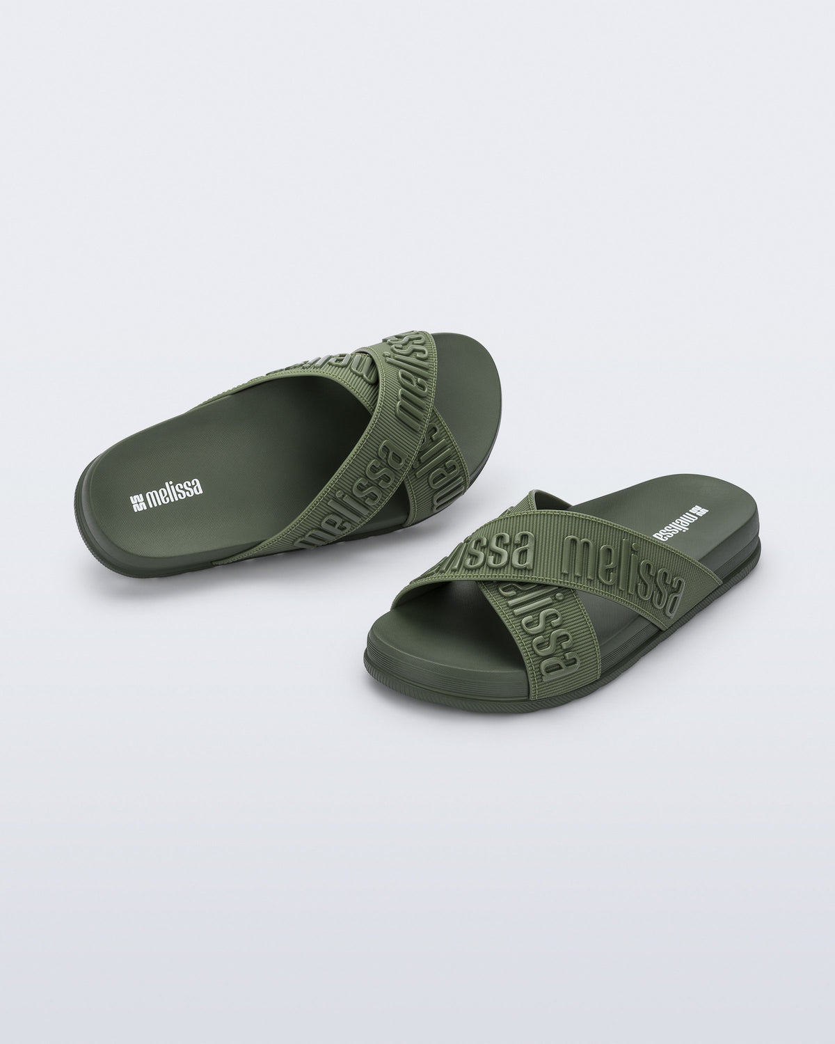 Angled view of a pair of Melissa Cozy slide sandals. The slide has two cross straps with Melissa logo embossed onto the straps. The color of the shoe is a dark green. 