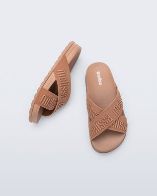 Top and side view of a pair of Melissa Cozy slide sandals. The slide has two cross straps with Melissa logo embossed onto the straps. The color of the shoe is brown.