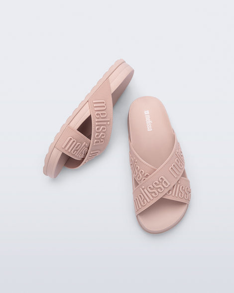 Top and side view of a pair of Melissa Cozy slide sandals. The slide has two cross straps with Melissa logo embossed onto the straps. The color of the shoe is light pink.