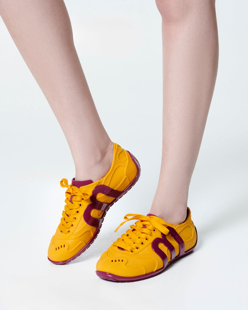 Model's legs wearing a pair of orange-yellow Motion sneakers with pink colored sole and M monogram