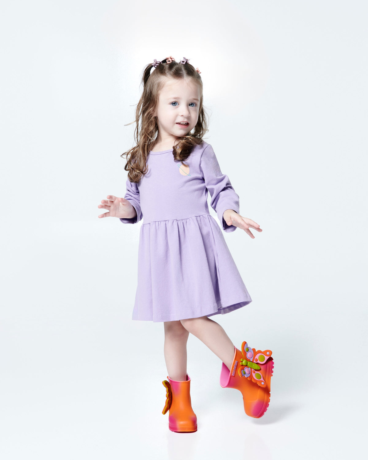 Child model in a lilac dress wearing a pair of orange with pink splotches Welly Wonderland baby boot with orange/green butterfly on side
