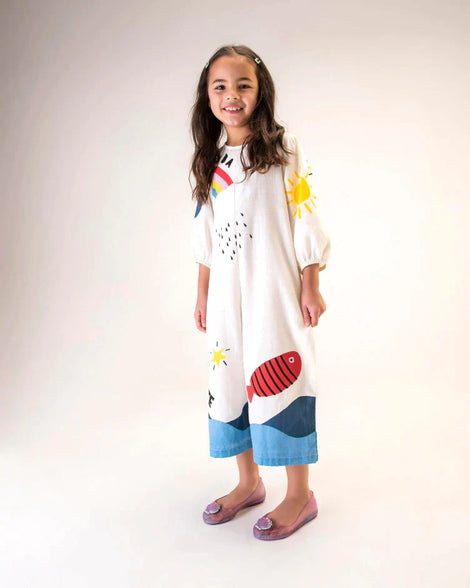 A young model posing for a picture in a white jumpsuit and a pair of purple Mini Melissa Sweet Love Princess flats, with a purple base, Ariel in script on the side, a sea shell detail on the toe, and a drawing of Ariel of The Little Mermaid on the sole.