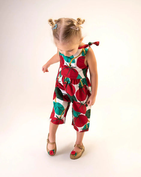 A young model in a patterned jumpsuit and a pair of gold/red Mini Melissa Sweet Love Princess flats with a gold base with Belle in script on the side, a top strap, a rose detail on the toe and a drawing of Belle of Beauty and the Beast on the insole.