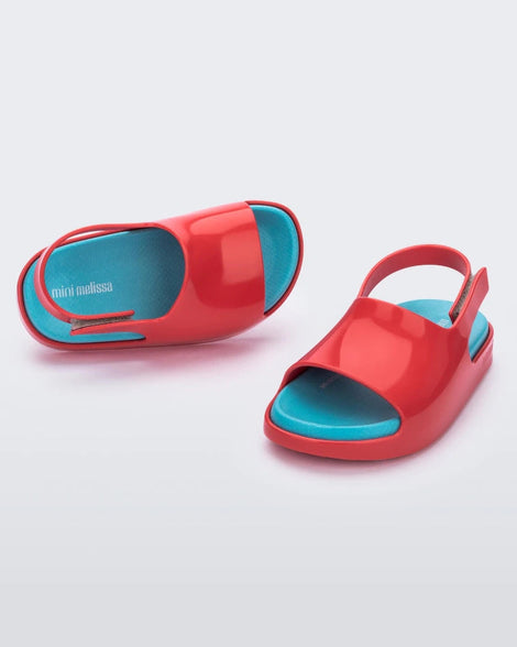 An angled front and top view of a pair of red Mini Melissa Cloud sandals with a blue insole, red base and back velcro strap.