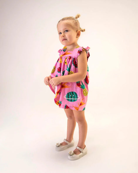 A young model in a pink dress, posing for a picture in a pair of Metallic White/Green Mini Melissa Sweet Love Princess flats with a metallic white base with Tiana in script on the side, a top strap, a flower detail on the toe and a drawing of Tiana of The Princess and The Frog on the insole.