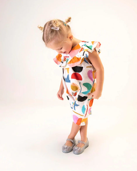 A young model in a white patterned dress and a pair of glitter blue/blue Mini Melissa Sweet Love Princess flats, with a blue glitter base, a top strap, Elsa in script on the side, a bow detail on the toe, and a drawing of Elsa of Frozen on the sole.