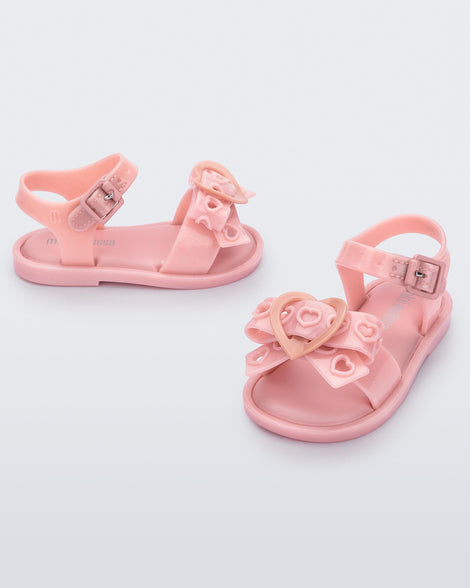 An angled front and side view of a pair of glitter pink Mini Melissa Mar Sandal Heart sandals with a glitter pink heart bow detail on the front strap and an ankle strap