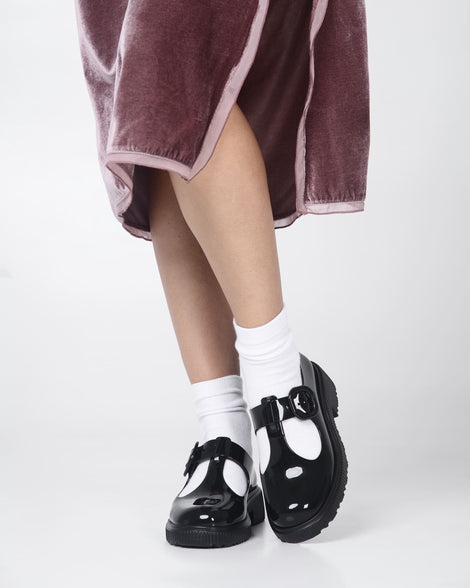 A model's legs wearing a skirt and a pair of black Melissa Jackie loafer with two cut outs and a buckle detail strap.