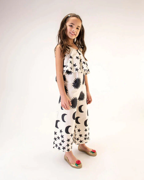 A model in a black and white patterned jumpsuit, wearing a pair of gold Mini Melissa Sweet Love Princess flats, with a gold base, Belle in script on the side, a rose detail on the toe, and a drawing of Belle of Beauty and the Beast on the sole.