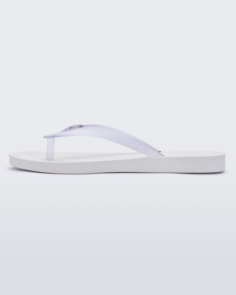 An inner side view of a white/clear Melissa Sun Venice flip flop with a white sole and clear straps with "melissa" on one strap.