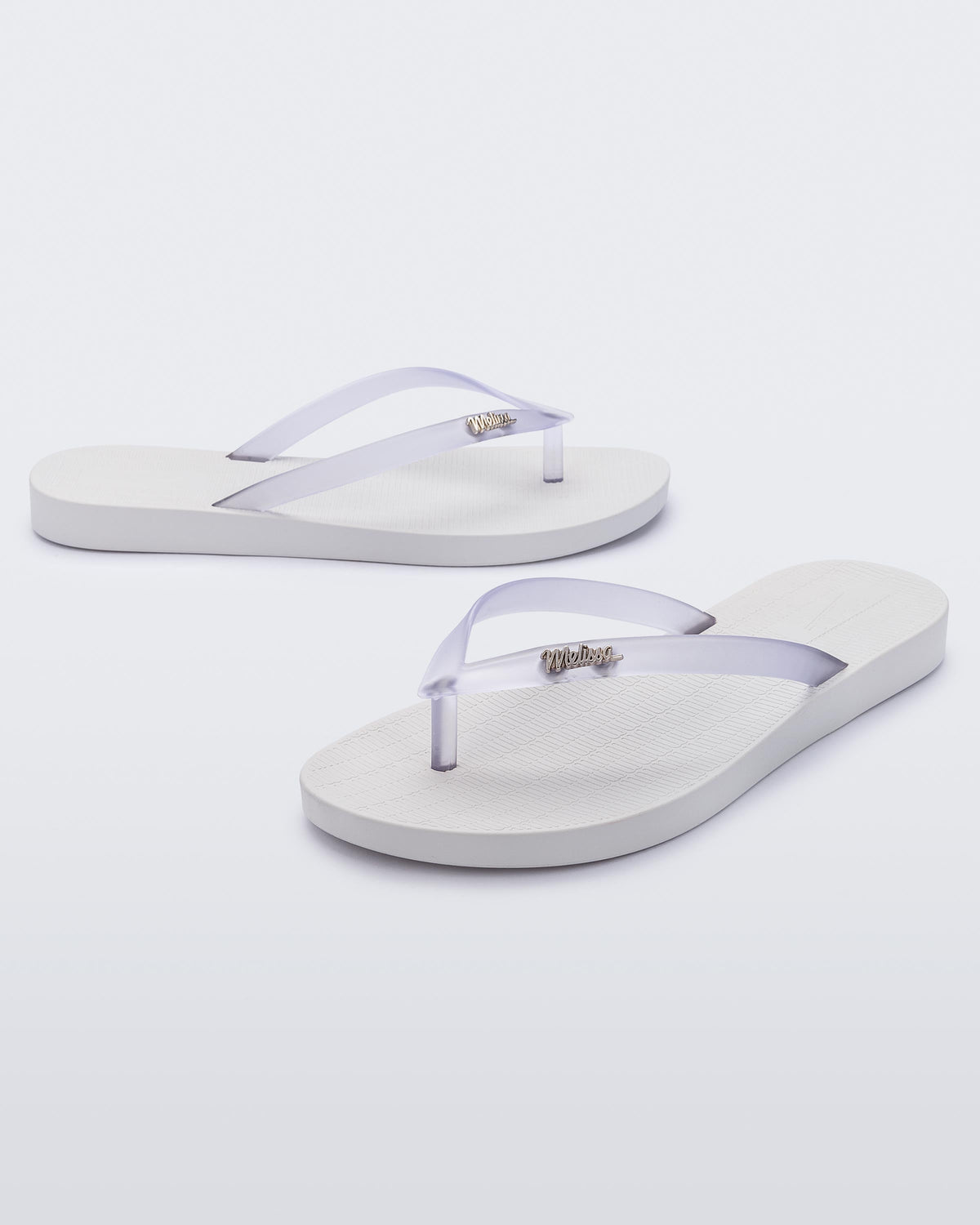 An angled front and side view of a pair of white/clear Melissa Sun Venice flip flops with a white sole and clear straps with "melissa" on one strap.