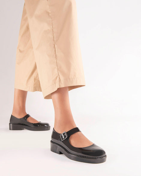 A model's legs wearing yellow pants and a pair of black Melissa Lola loafers with a rounded toe and a black strap and buckle.