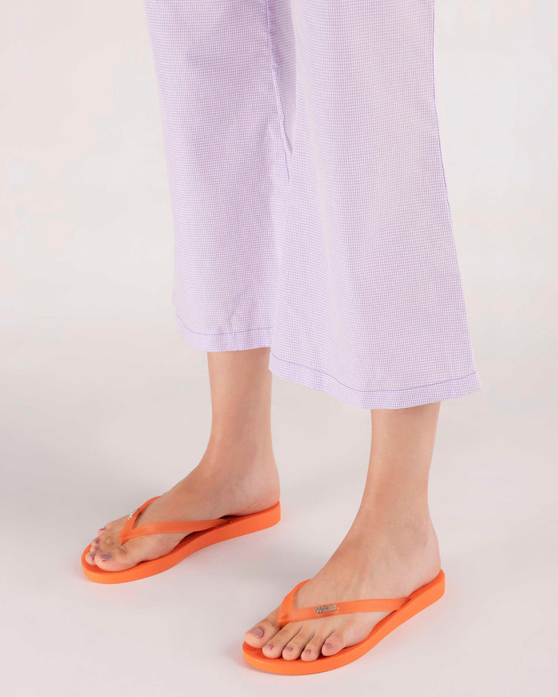 A model's legs in purple gingham pants, wearing a pair of orange Melissa Sun Venice flip flops with "melissa" on the strap.