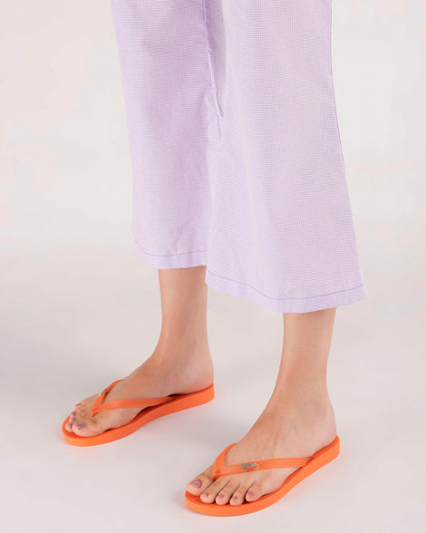 A model's legs in purple gingham pants, wearing a pair of orange Melissa Sun Venice flip flops with 