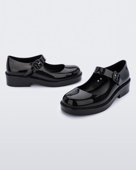 An angled front and side view of a pair of black Melissa Lola loafers with a rounded toe and a black strap and buckle.